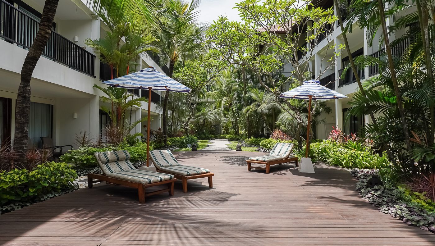 Garden walkway with loungers and parasols between resort buildings at Away Bali Legian Camakila, Legian
