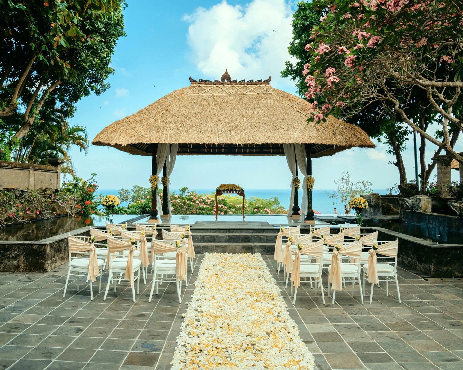 A thatched Balinese bale set on a water platform above the ocean at Bale Kencana, AYANA, with a stone terrace and yellow-petal aisle
