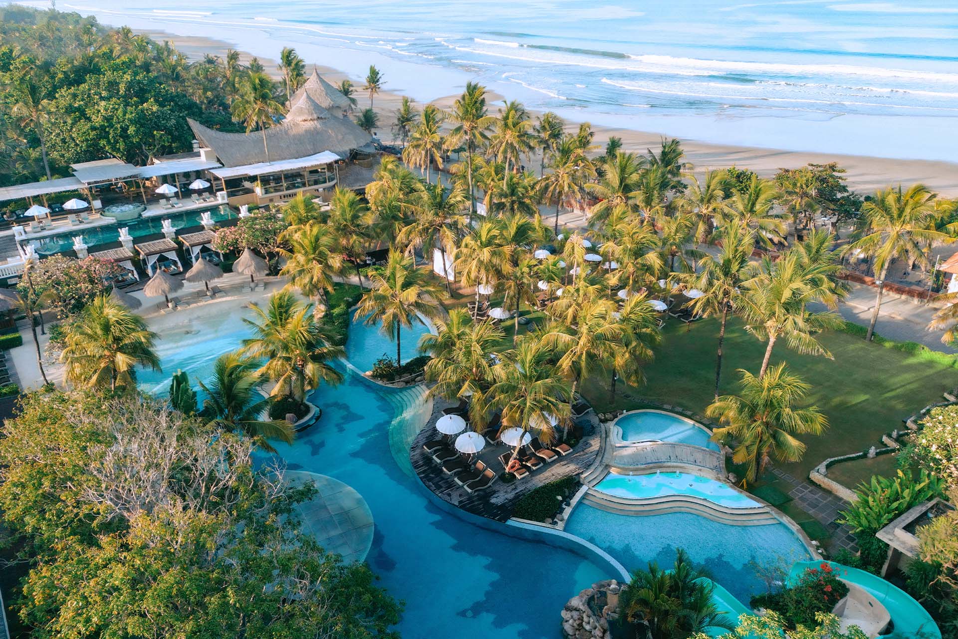 Aerial view of the lagoon pools, palm gardens, and Legian Beach at Bali Mandira Beach Resort & Spa