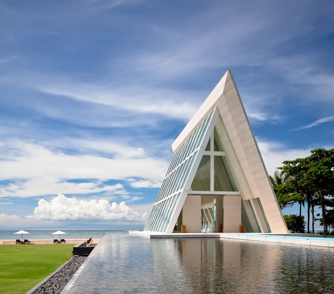 The triangular glass Infinity Chapel at Conrad Bali appearing to float on reflection pools with the beach and Indian Ocean behind