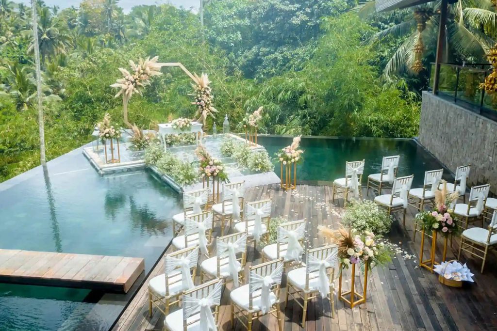 A water wedding set up on the glass stage over the infinity pool at Kaamala Resort Ubud, with gold chairs on the deck and forest beyond