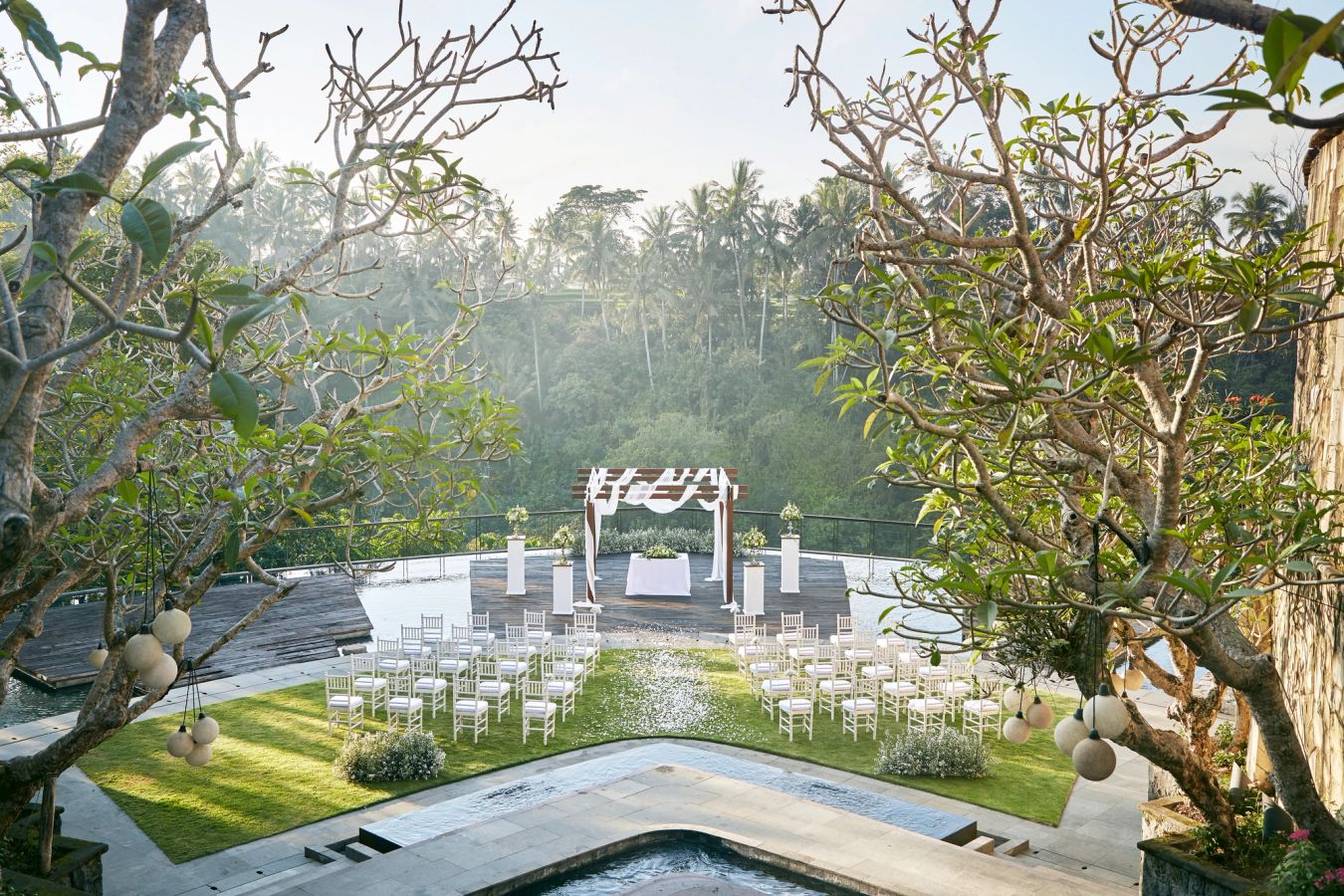 An open-air wedding ceremony set up on a deck over the valley at Kamandalu Ubud, framed by frangipani trees with rice terraces and palms beyond