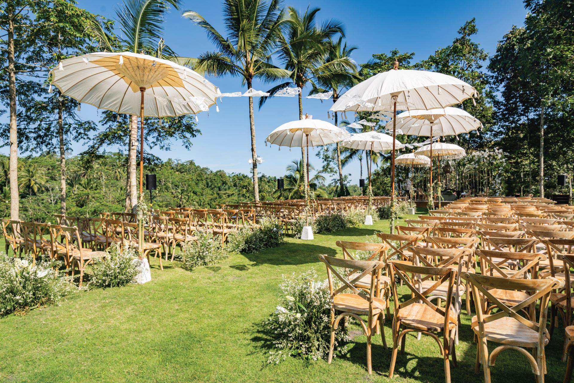 A garden ceremony on the wedding lawn at Padma Resort Ubud, with white Balinese ceremonial umbrellas, wooden chairs, and jungle forest behind