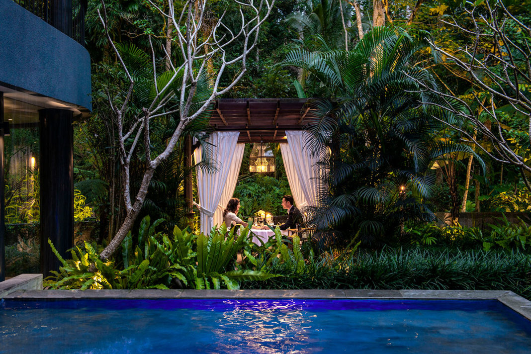A couple at a private candlelit dinner in a lantern-lit pavilion beside a pool, surrounded by rainforest at Samsara Ubud