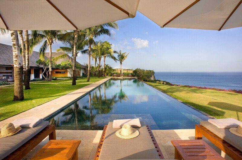The long infinity pool and lawn at The Istana in Uluwatu, framed by palms and daybeds with the Indian Ocean beyond the cliff