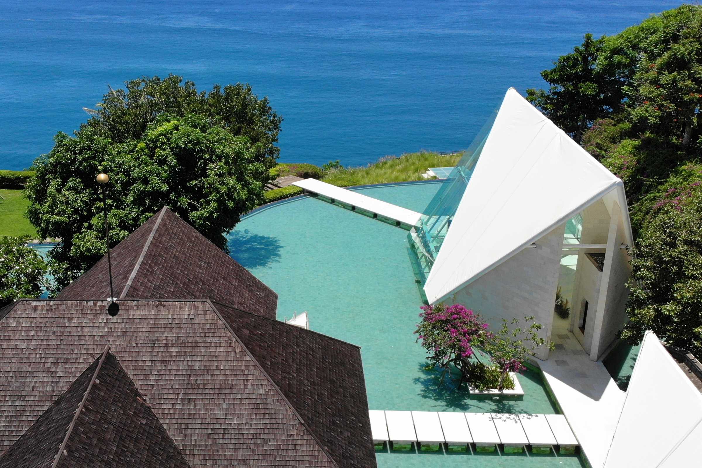 Aerial view of the Tirtha Uluwatu glass chapel over water on the Uluwatu clifftop, with the Indian Ocean behind