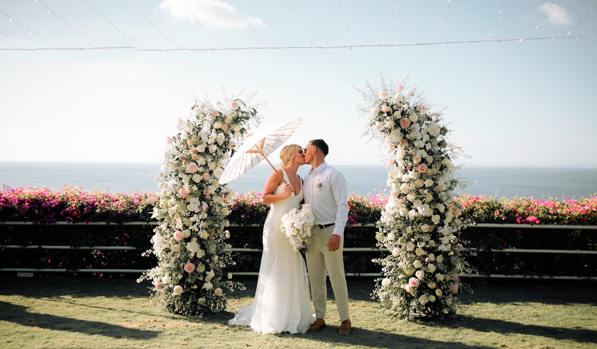 A couple kissing under a parasol beneath a floral ceremony arch on the clifftop lawn at Uluwatu Cottages, ocean and bougainvillea behind