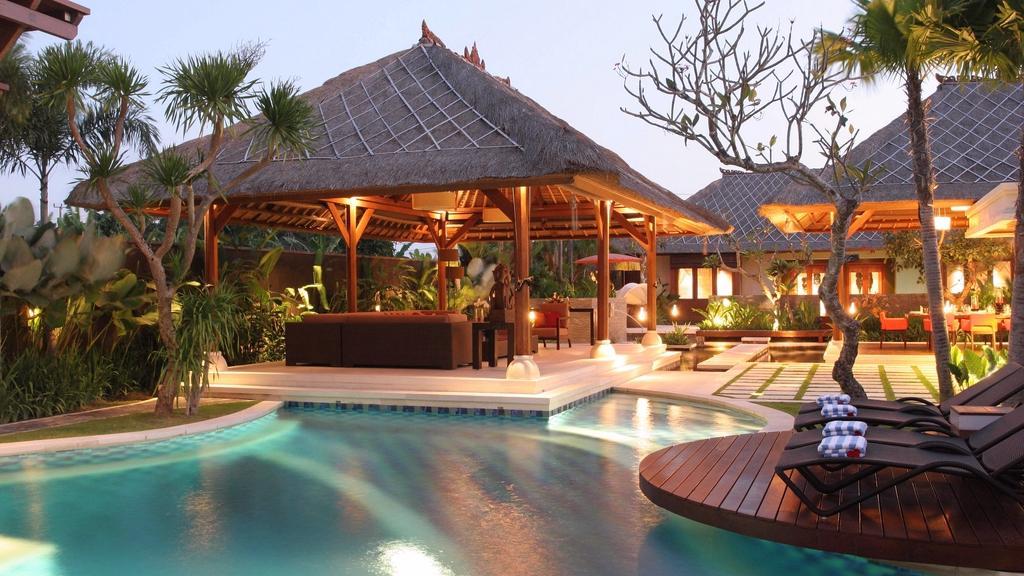 The free-form pool at Villa Asta at dusk, with the thatched-roof pavilion and sun loungers lit for the evening