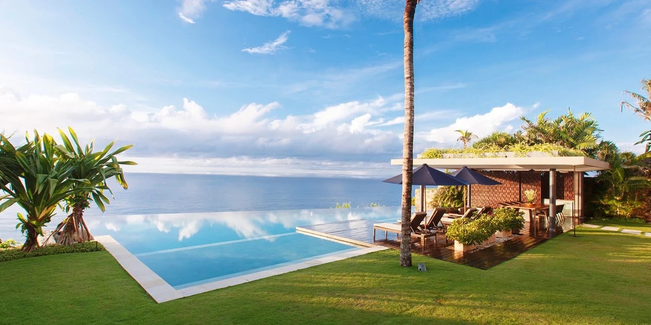 Cliff-edge infinity pool at Villa Jamadara, The Ungasan, reflecting the sky with a tall palm and the Indian Ocean beyond