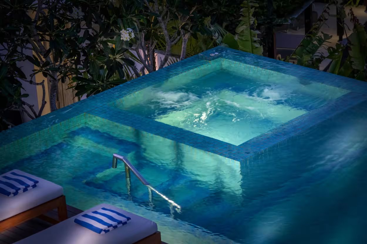 The blue-tiled pool and raised jacuzzi at Villa KaliBali lit at night, with sun loungers and frangipani trees on the Pecatu hillside in Uluwatu