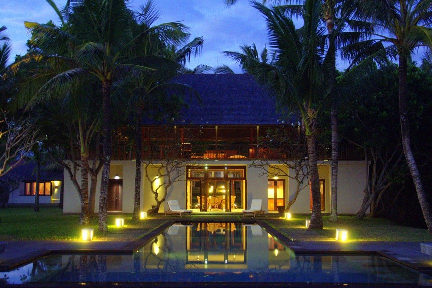 Villa Lalang's pool and two-storey villa lit up at twilight, palm trees framing the Balian coastline in West Bali