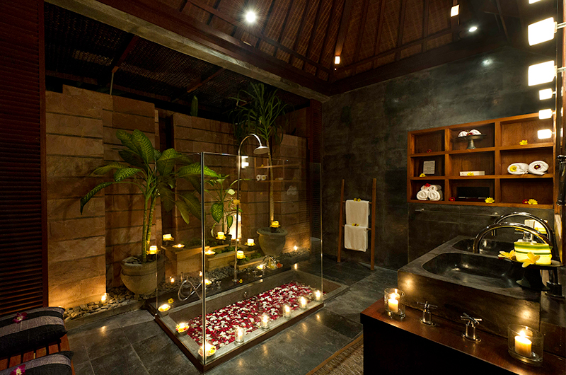 Candlelit stone bathroom with a petal-strewn bath under a Joglo roof at Villa Maya, Majapahit Beach Villas