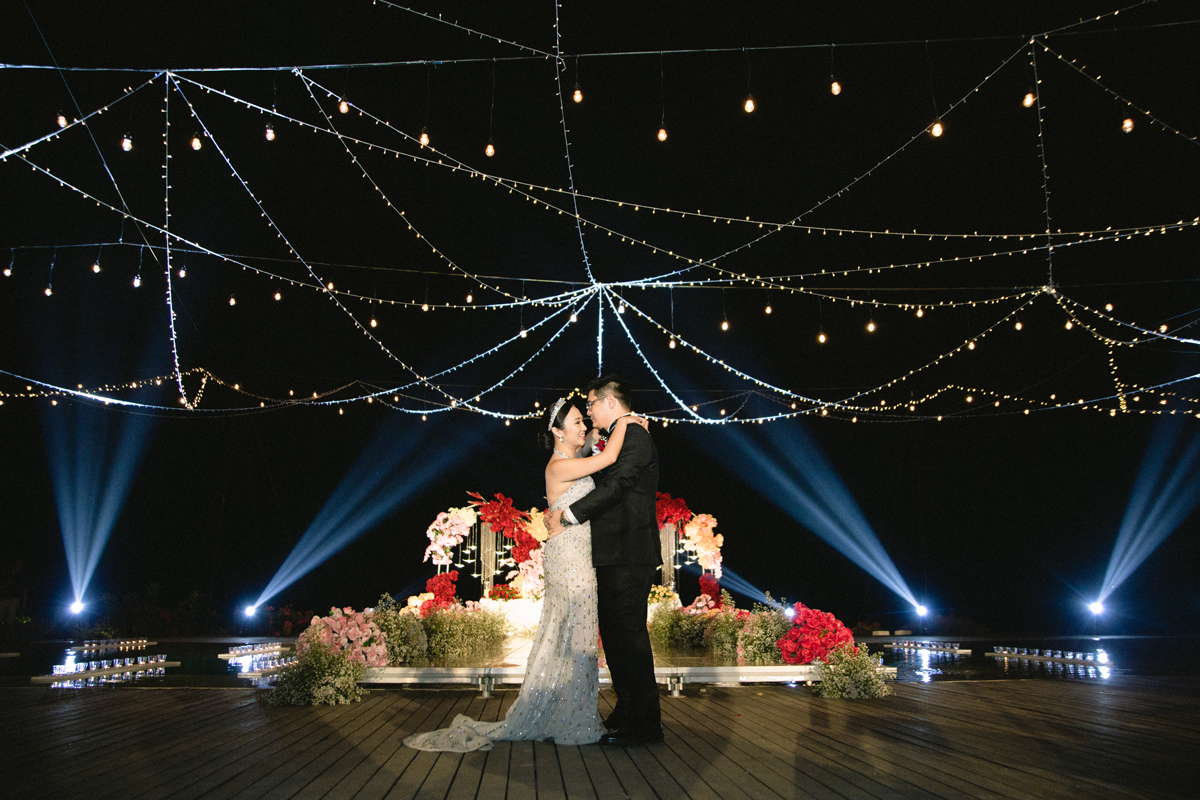 An evening wedding on the cliff-edge deck at Wonderland Uluwatu, a couple under strings of fairy lights with a floral stage and the dark ocean behind